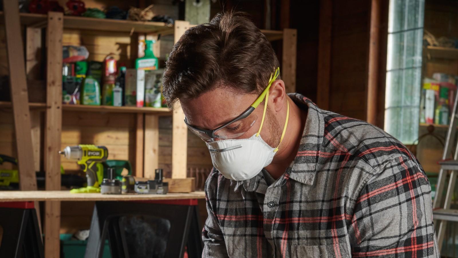 Muž pracujúci v dielni má nasadené Ryobi ochranné okuliare a respirátor.